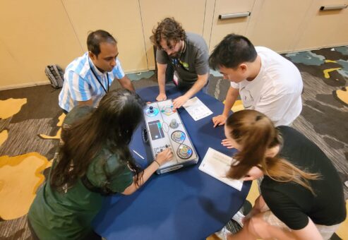 Five participants leaning over a Team Synergy retro gaming console, strategizing and solving mini-game challenges during a team building activity.
