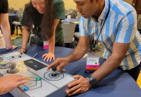 Team member enthusiastically interacting with the Team Synergy console while teammates look on and laugh during a fun team building challenge.