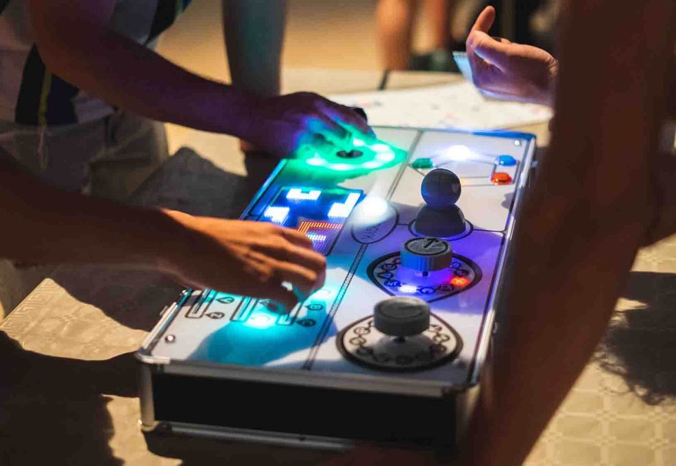 Team members collaborate on a brightly lit retro gaming console, using buttons, joysticks, and sliders during the Team Synergy team building event.