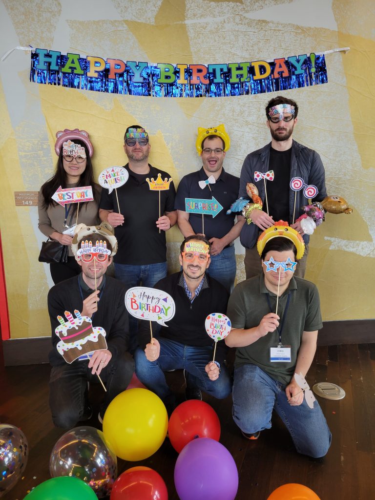 Group of participants posing with fun birthday-themed props and wearing party accessories in front of a 'Happy Birthday' banner during the Birthday Kits activity at a corporate team building event, called 'The Donation Station'.