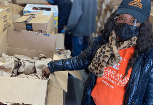 Volunteer wearing a mask and orange shirt organizing brown paper bags in a donation center filled with boxes.