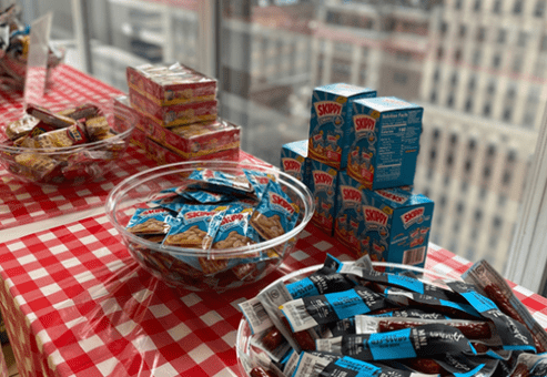 Snacks and food items, including Skippy peanut butter packs and granola bars, arranged on a red checkered table at a corporate charitable team building event.