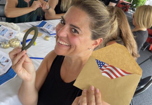 Smiling woman holding an envelope with an American flag and a small parcord bracelet during a corporate team building event, with other participants working in the background.