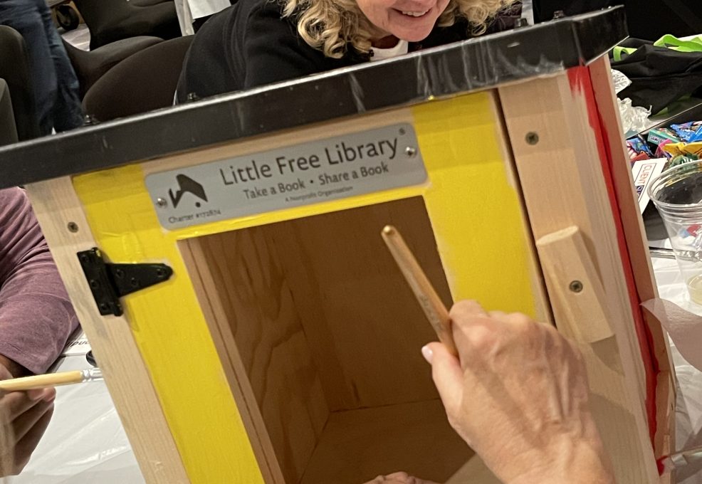 A person paints the outer frame of a wooden Little Free Library box in bright yellow while another person watches. The box is mounted on the table and other materials for decorating the box are scattered in the background.