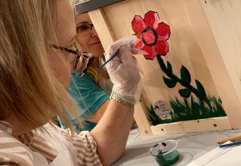 Participant painting a red flower on a wooden Little Free Library as part of the Little Team Library team building event, where teams assemble and decorate book exchange boxes for the community.