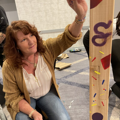A team member paints a Little Free Library® post with colorful designs during the Little Team Library charitable team building program, promoting literacy and teamwork. thumbnail