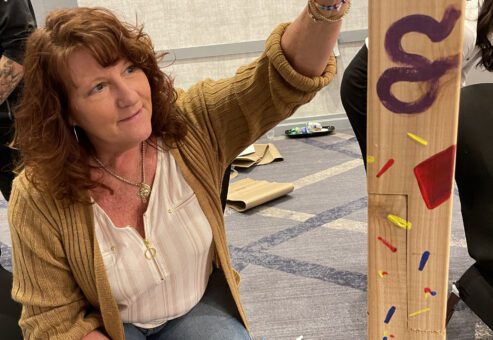 A team member paints a Little Free Library® post with colorful designs during the Little Team Library charitable team building program, promoting literacy and teamwork.