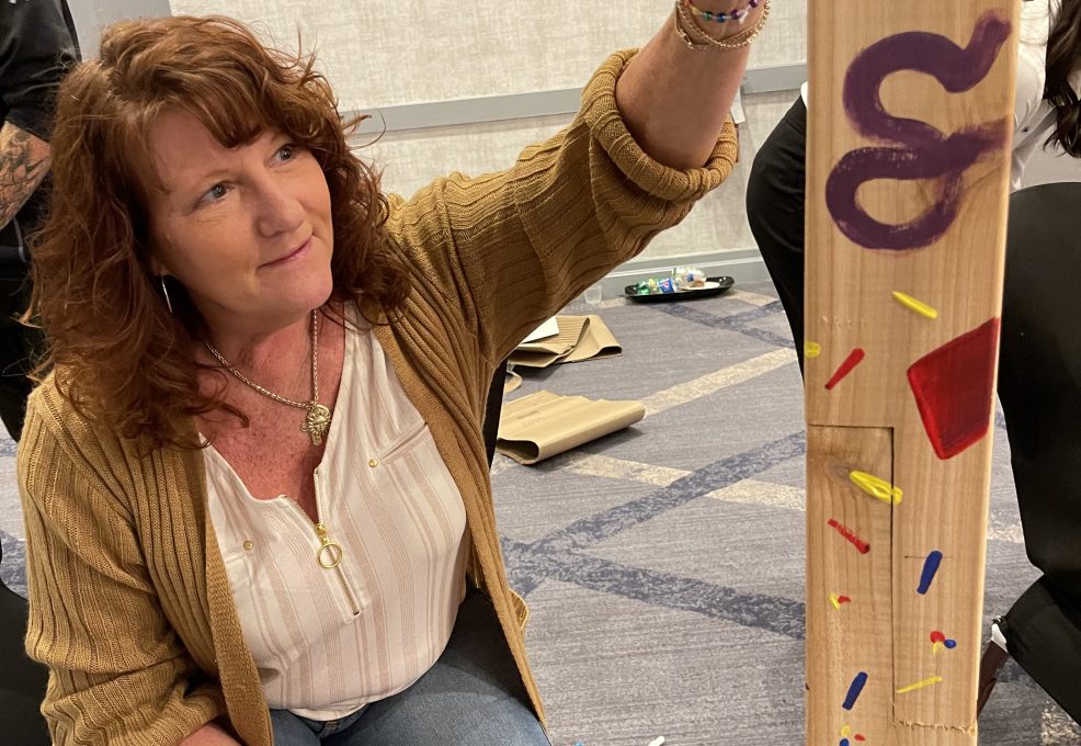 A team member paints a Little Free Library® post with colorful designs during the Little Team Library charitable team building program, promoting literacy and teamwork.