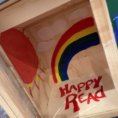 Interior of a decorated Little Free Library featuring a painted rainbow, sun, clouds, and the words 'Happy Read' as part of the Little Team Library team building event. thumbnail