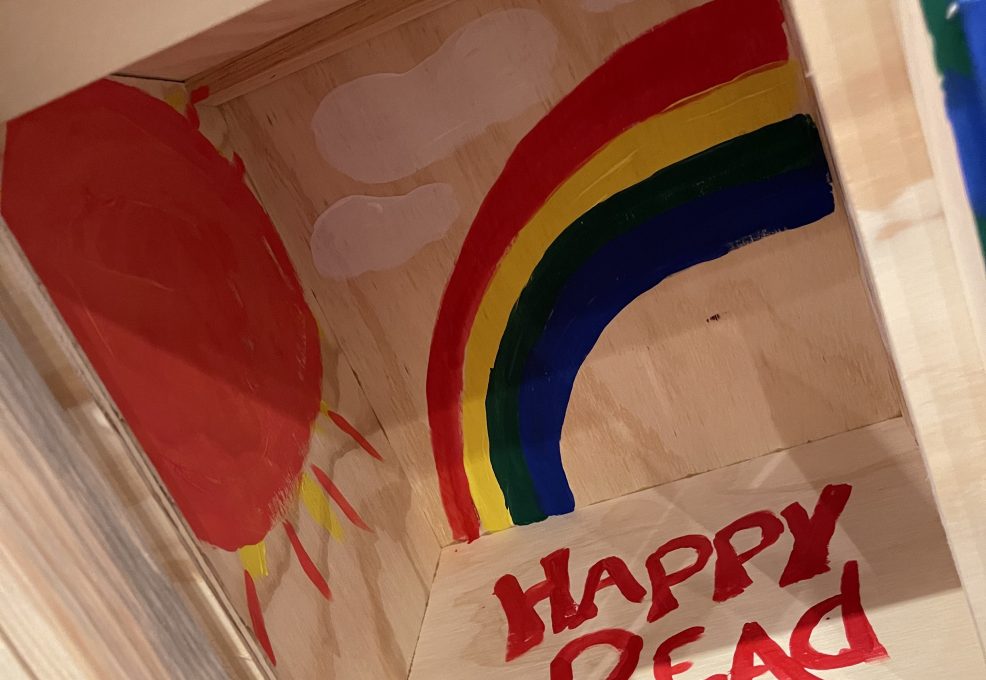 Interior of a decorated Little Free Library featuring a painted rainbow, sun, clouds, and the words 'Happy Read' as part of the Little Team Library team building event.