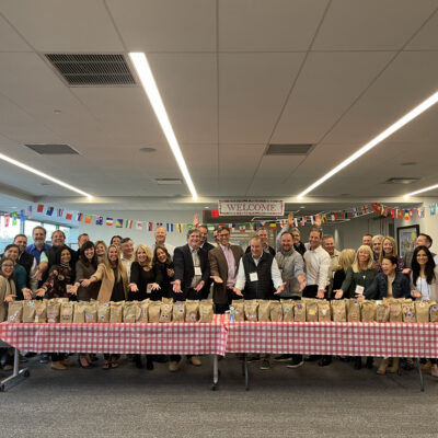 Large group of people posing behind a long table filled with decorated brown paper bags during a corporate giving team building event, with international flags hanging in the background. thumbnail