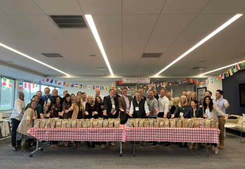 Large group of people posing behind a long table filled with decorated brown paper bags during a corporate giving team building event, with international flags hanging in the background.