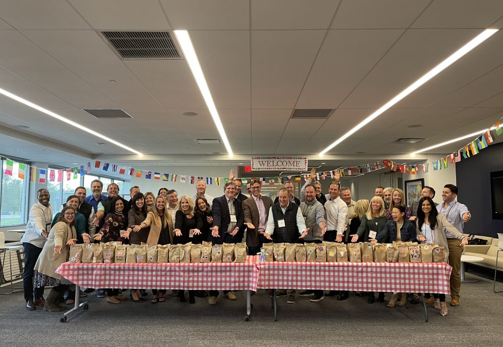 Large group of people posing behind a long table filled with decorated brown paper bags during a corporate giving team building event, with international flags hanging in the background.