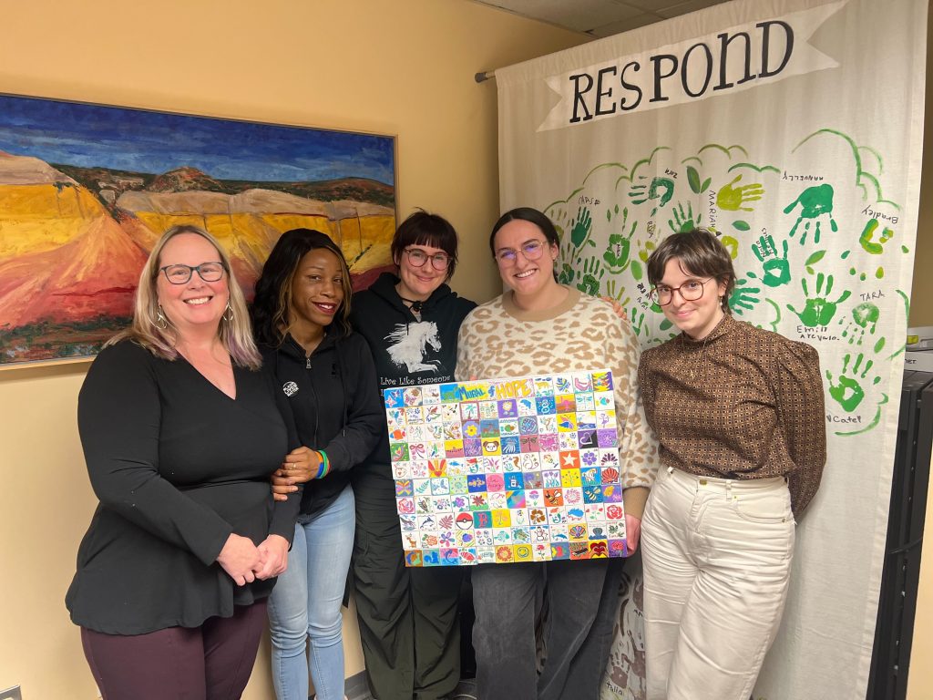 Group of participants smiling and holding a colorful mini-mural made of small painted tiles during the Make-A-Mini-Mural activity at a corporate charitable team building event called 'the Donation Station'.