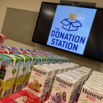 Juice boxes stacked on a table in front of a screen displaying 'The Donation Station' logo during a corporate charitable team building event. thumbnail