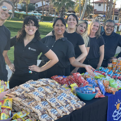 Group of facilitators smiling behind a table filled with snacks and drinks at an outdoor corporate charitable team building event. thumbnail