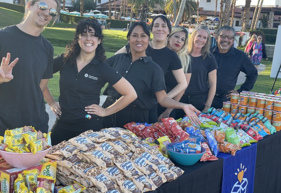 Group of facilitators smiling behind a table filled with snacks and drinks at an outdoor corporate charitable team building event.