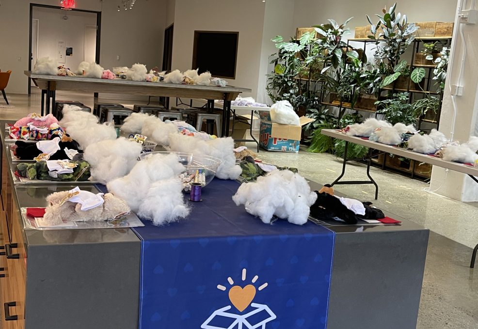 Donation Station setup with tables full of kit materials, including white packing material and small items for assembling donation kits. A banner with 'The Donation Station' is displayed in a brightly lit room, ready for a corporate charitable team building event.