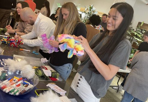 Participants at a Donation Station event assembling colorful donation kits, with supplies like markers and stuffing on the table. A woman in the foreground smiles as she works on a stuffed toy.