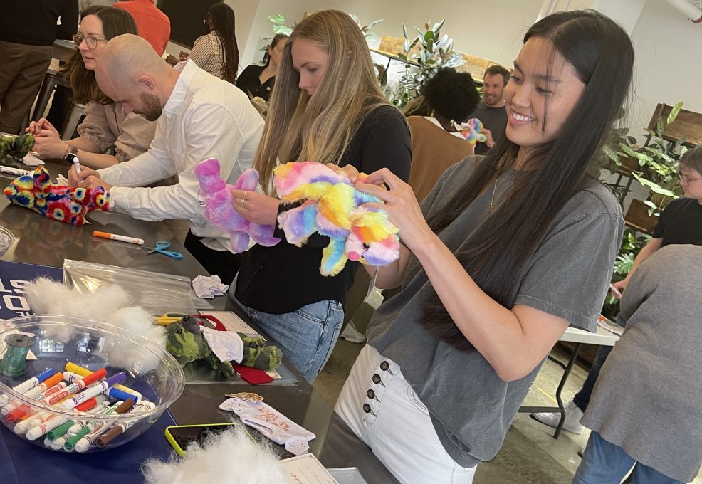 Participants at a Donation Station event assembling colorful donation kits, with supplies like markers and stuffing on the table. A woman in the foreground smiles as she works on a stuffed toy.