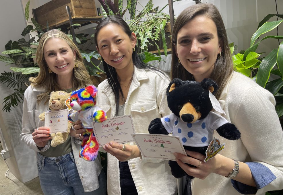 Three women smiling and holding colorful stuffed animals and birth certificates they created during a corporate charitable team building event called The Donation Station.