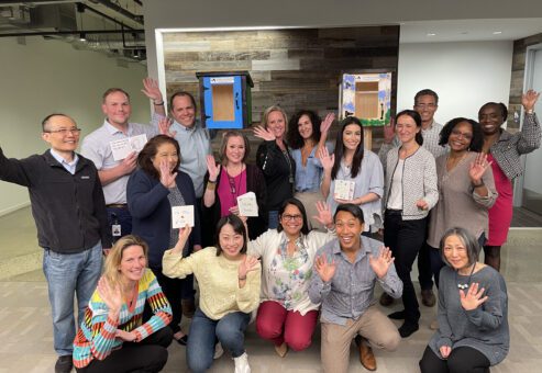 Group of participants smiling and waving in front of two completed Little Free Libraries during the Little Team Library team building event.
