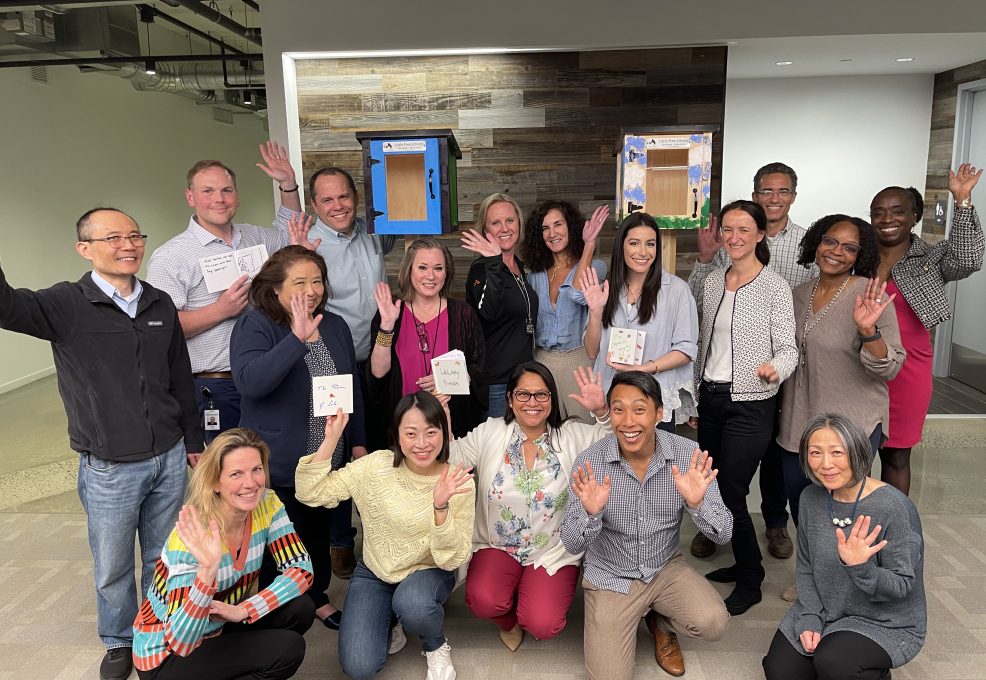Group of participants smiling and waving in front of two completed Little Free Libraries during the Little Team Library team building event.