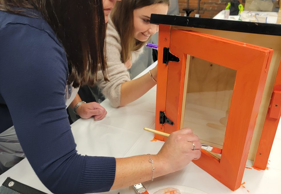 Participants focus on painting a Little Free Library door with bright orange during a collaborative team building event. The hands-on activity fosters creativity and teamwork as teams work together to build and decorate the library for community use.