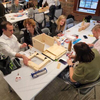 Teams collaborate to assemble a Little Free Library during a hands-on team building event. Participants work together to construct the wooden structure, following instructions and dividing tasks to create a functional and beautifully decorated library for the community. thumbnail