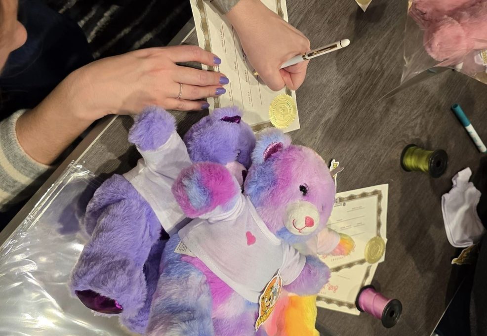 A person's hand is seen writing on a certificate next to colorful stuffed animals and spools of thread on a table.