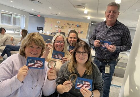 A group of five people holds small American flags and bracelets, smiling in a bright, modern indoor space.