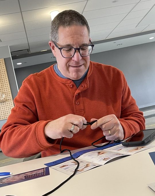 A man in an orange sweater is focused on connecting a cable while sitting at a table with documents and a pen.