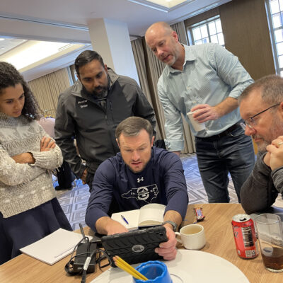 A group of five team members gathered around a tablet, focusing on completing a task during the Wheels Around the World team building event. thumbnail