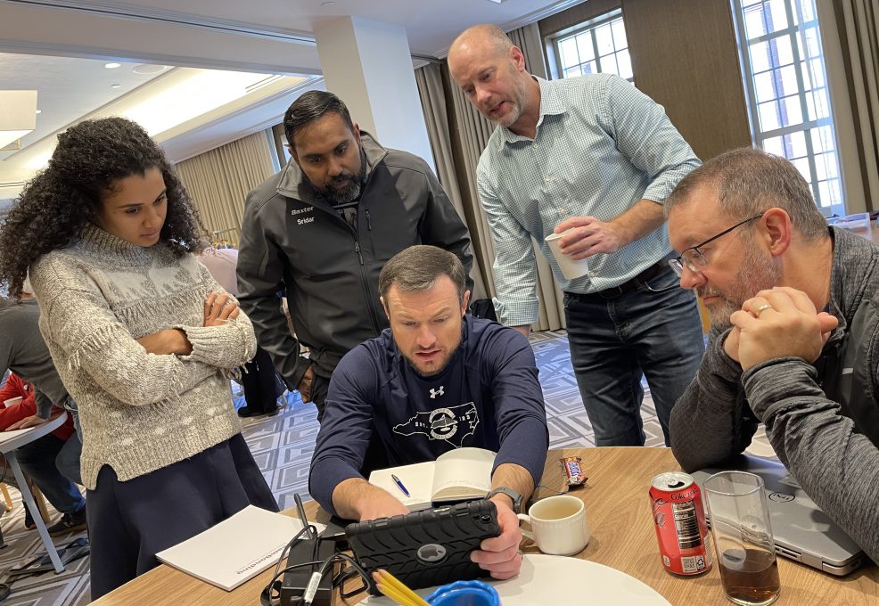 A group of five team members gathered around a tablet, focusing on completing a task during the Wheels Around the World team building event.