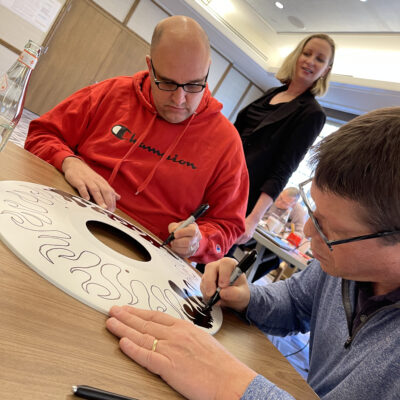 Two participants focus on decorating a wheelchair safety cover with intricate black designs during the Wheels Around the World team building event. thumbnail