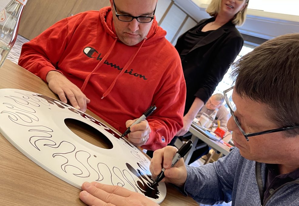 Two participants focus on decorating a wheelchair safety cover with intricate black designs during the Wheels Around the World team building event.