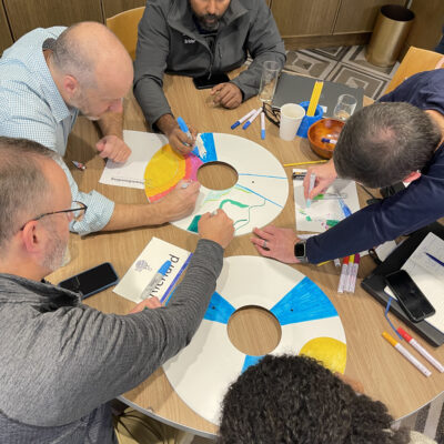 Team members gathered around a table, decorating wheel safety covers with colorful designs as part of the Wheels Around the World team building event. thumbnail
