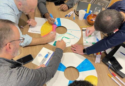 Team members gathered around a table, decorating wheel safety covers with colorful designs as part of the Wheels Around the World team building event.