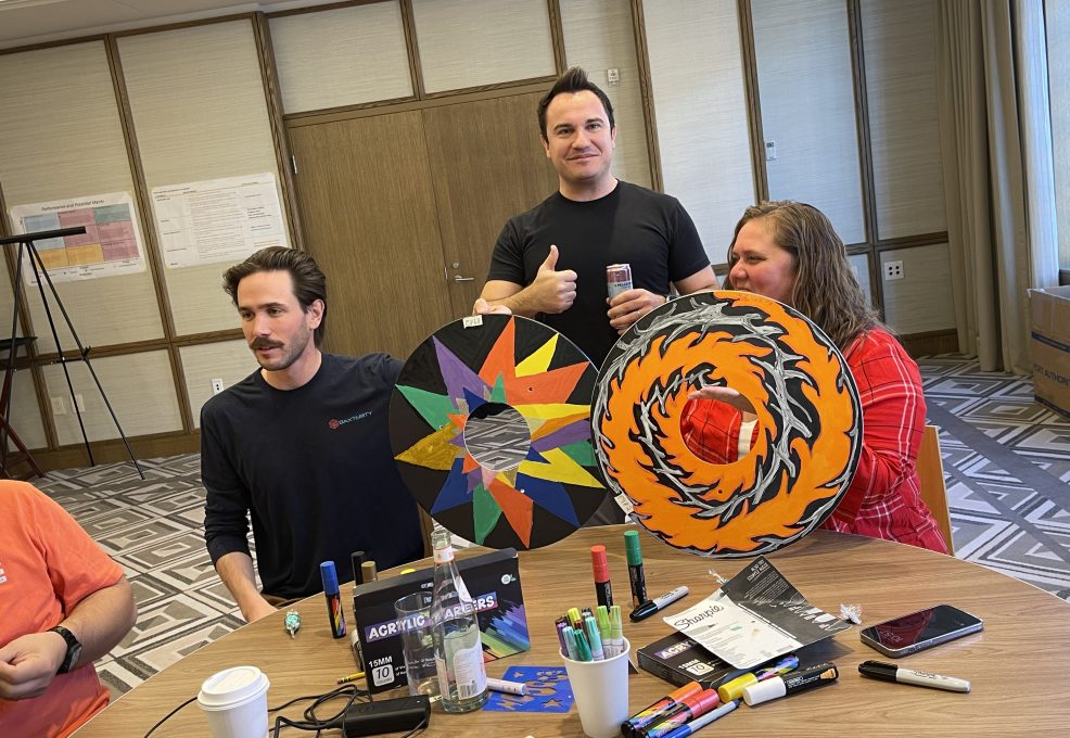 Three participants in the Wheels Around the World team building event display their creatively decorated wheelchair safety covers. The covers feature vibrant designs, showcasing the team's artistic contribution to the charitable project.