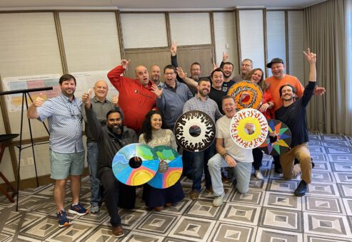 A group of enthusiastic participants proudly displaying personalized wheelchair safety covers they decorated during the Wheels Around the World team building event. The decorated wheels feature colorful designs, and the group celebrates their contribution to making a positive impact.