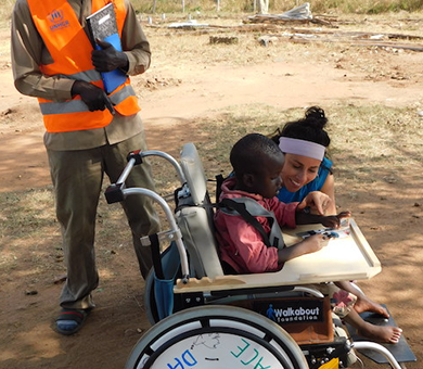 A young boy receiving a decorated wheelchair during the Wheels Around the World team building event, accompanied by two adults who are assisting him outdoors.