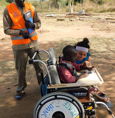 A young boy receiving a decorated wheelchair during the Wheels Around the World team building event, accompanied by two adults who are assisting him outdoors. thumbnail