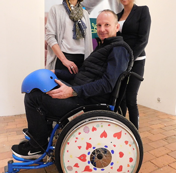 Two team members pose next to a man seated in a decorated wheelchair featuring heart designs on the wheel cover, created during the Wheels Around the World team building event.
