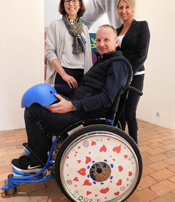 Two team members pose next to a man seated in a decorated wheelchair featuring heart designs on the wheel cover, created during the Wheels Around the World team building event. thumbnail