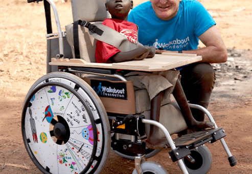 A young boy sits in a newly donated wheelchair, customized with a decorated wheel safety cover, next to a smiling volunteer from the Walkabout Foundation during the Wheels Around the World team building event.