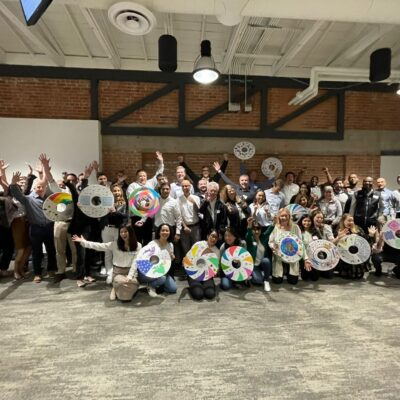 A large group of participants proudly display their decorated wheel safety covers during the Wheels Around the World team building event, celebrating their contribution to providing wheelchairs for individuals in need. thumbnail