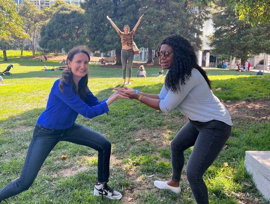 Two women participate in a playful team building activity in a park. They are posing while balancing something in their hands, with a third person standing in the background with arms raised in celebration.