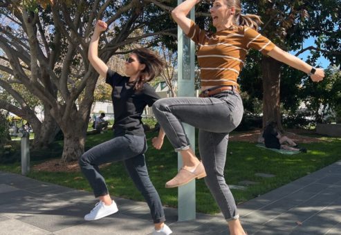 Two women energetically jump in the air during a team building activity outdoors, with trees in the background. Both are striking playful poses mid-jump, adding a sense of fun and excitement to the moment.