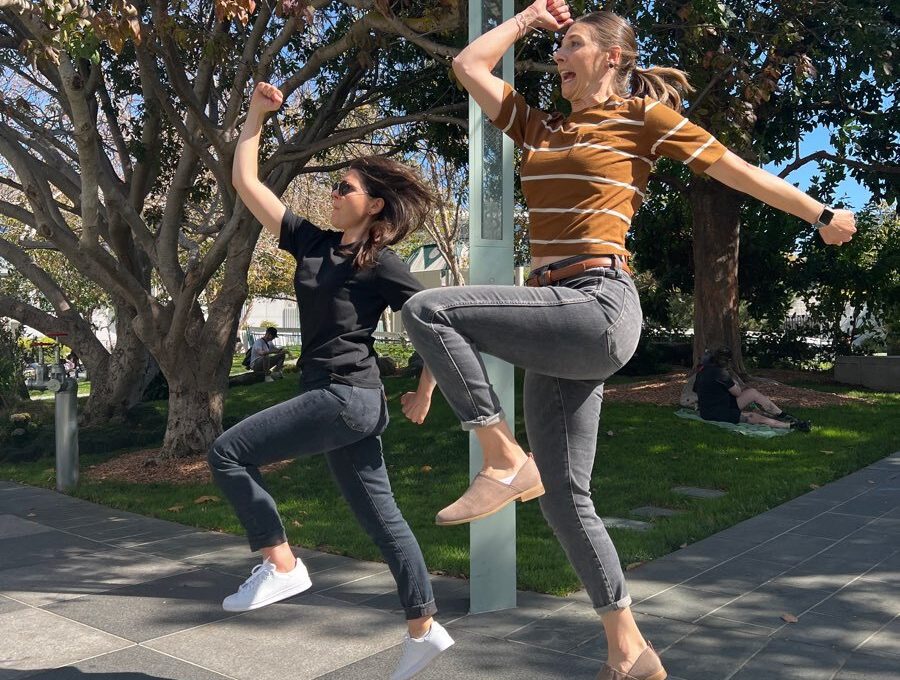 Two women energetically jump in the air during a team building activity outdoors, with trees in the background. Both are striking playful poses mid-jump, adding a sense of fun and excitement to the moment.