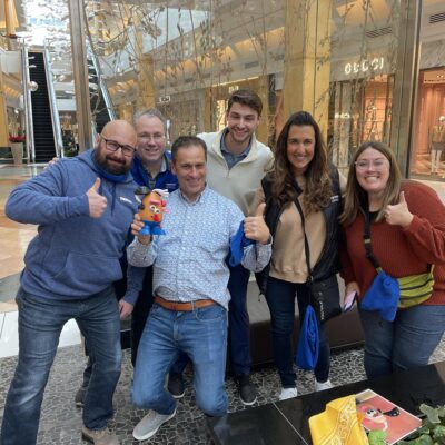Group of six participants smiling and giving thumbs up during a team building event in a shopping mall, holding a Mr. Potato Head toy. thumbnail